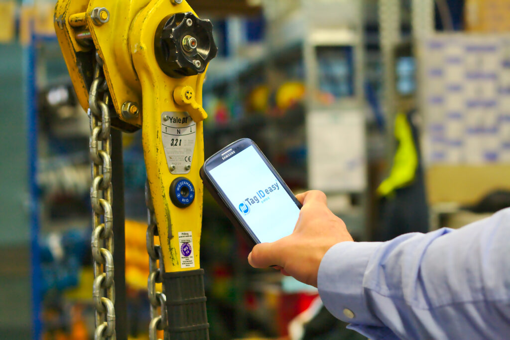 Mann scannt mit Smartphone einen RFID Tag, um das Betriebsmittel für eine UVV-Prüfung zu identifizieren, um die Arbeitssicherheit zu gewährleisten.